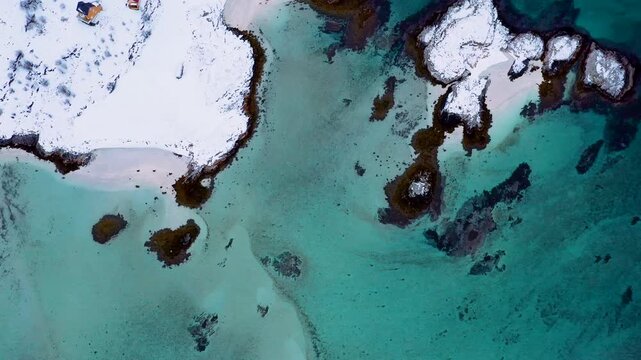 Drohnenaufnahme einer einzelnen isolierten H&uuml;tte auf schneebedeckter Insel mit t&uuml;rkisfarbenem arktischen Wasser, Nordnorwegen 4K
