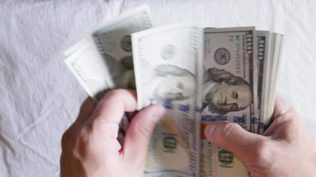 Adult man counts a stack of US dollar bills indoors on a light fabric surface. Scene conveys feelings of wealth and financial abundance.