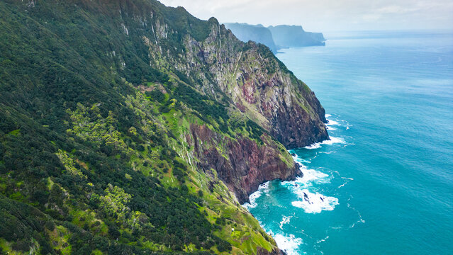 Dramatic aerial view of Madeira's lush green cliffs plunging into the turquoise Atlantic, with waves crashing against the rugged coastline under an overcast sky. Vereda do Larano