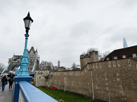 Blue lamppost and pedestrian view toward Tower Bridge with Tower of London walls on a cloudy day in London, UK