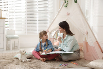 Mother and her daughter drawing in sketchbook together near toy wigwam at home, space for text © New Africa