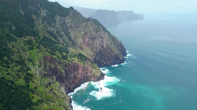 Dramatic aerial view of Madeira's lush green cliffs plunging into the turquoise Atlantic, with waves crashing against the rugged coastline under an overcast sky. Vereda do Larano