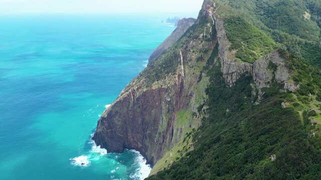Dramatic aerial view of Madeira's lush green cliffs plunging into the turquoise Atlantic, with waves crashing against the rugged coastline under an overcast sky. Vereda do Larano