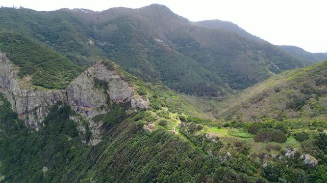 Aerial view of Madeira's lush mountains and valleys. Green slopes, a distinctive rocky peak, and distant village hint at island's natural beauty and hiking trails. Vereda do Larano