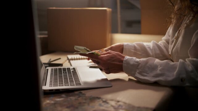 Female Handling Cash And Orders, Female Worker Preparing Packages While Reviewing Mobile App And Notes