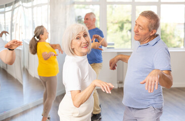 Fototapeta premium Cheerful enthusiastic senior lady enjoying active dancing paired with male partner during group training in dance studio, practicing playful twist..
