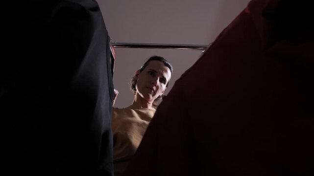 Woman standing in front of an open closet or wardrobe, looking up at clothes hanging on a rail with a frustrated expression, experiencing difficulty making a choice for her outfit