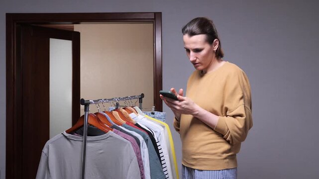 Woman standing next to a clothing rack, browsing and selecting garments using a smartphone, focusing on sustainable fashion choices or decluttering an existing wardrobe