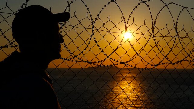 Man observing sunset behind barbed wire fence. Solitary silhouetted male standing against dramatic sunset, gazing through barbed wire fence with expansive ocean horizon, symbolizing hope and isolation
