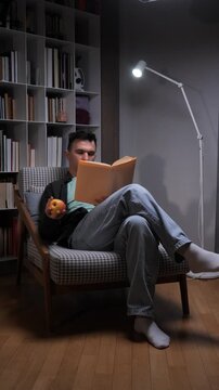 Vertical video man relaxing in an armchair, reading a book under the soft light of a floor lamp, holding an apple, surrounded by bookshelves in a cozy home environment