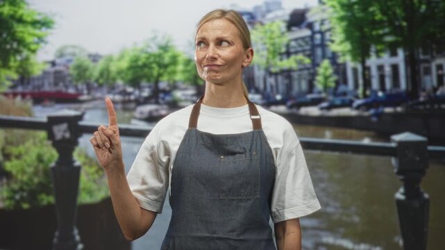Woman in apron points finger on street by a canal and bridge, slight smirk and direct pose; practical service skepticism.