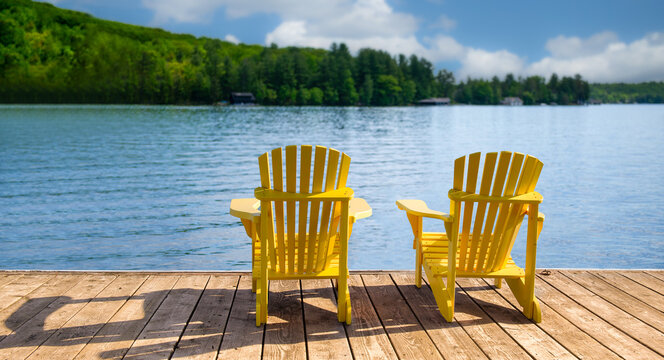 Rear view of two bright yellow Adirondack chairs on a weathered dock in Muskoka Ontario Canada, calm blue lake and forested hills, cottage country, no people, copy space.