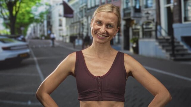 Woman midriff posing hands on hips on an urban street with row houses and parked car, smiling and looking aside; confidence wellness.