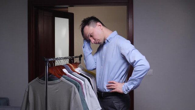 Young man standing in front of a wardrobe rack in a room, contemplating different clothing options and experiencing a dilemma choosing an outfit for the day