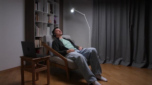 Man finding comfort and relaxation at home, sitting in a plaid armchair with headphones on, enjoying audio content from his laptop in a cozy evening setting
