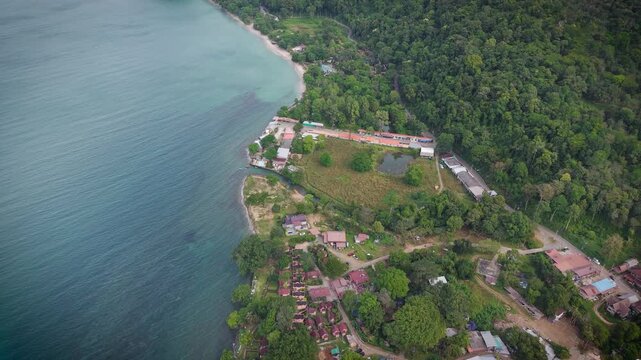 Aerial drone view of tropical beach, sea and Thai houses surrounded by lush forest. Scenic coastal landscape in Thailand with turquoise water and traditional buildings from above.