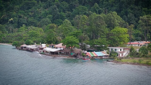 Aerial drone view of tropical beach, sea and Thai houses surrounded by lush forest. Scenic coastal landscape in Thailand with turquoise water and traditional buildings from above.