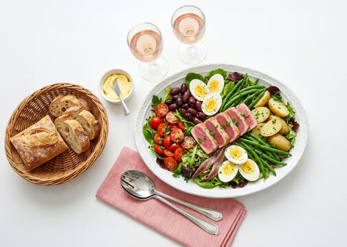 Ni&ccedil;oise salad with seared tuna, eggs, cherry tomatoes, potatoes, accompanied by a basket of baguette bread and ros&eacute; wine on a white background.