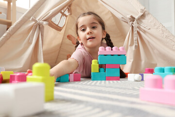 Little girl playing with building blocks near toy wigwam in playroom, low angle view © New Africa