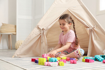 Little girl playing with building blocks near toy wigwam in playroom, space for text © New Africa