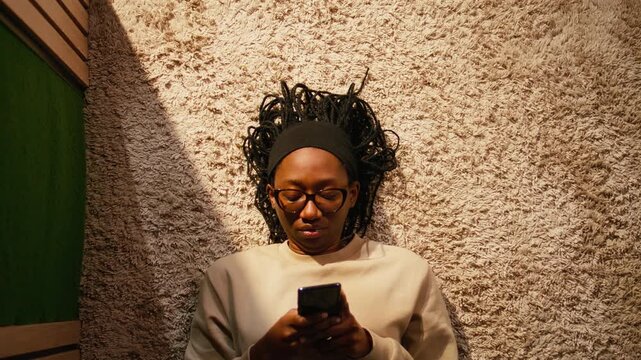 African American woman lying on the floor in cozy modern hallway, focused on smartphone while browsing apps and scrolling social media. Peaceful indoor scene with warm interior light.