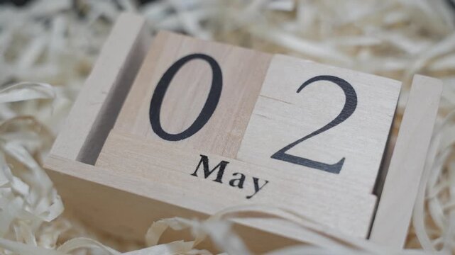 Wooden calendar blocks switched to May 2 resting on straw filler. Soft close-up shot with natural tones emphasizes seasonal transition and time passing.