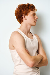 Obraz premium Young man with red hair and fair skin standing with arms crossed in white tank top against plain background. Profile portrait showing serious expression, casual style and youth.
