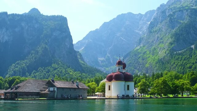 Cinematic video footage of the red-domed St. Bartholoma pilgrimage church on the shore of Konigsee lake, surrounded by high Alpine mountains in Berchtesgaden, Bavaria, Southern Germany.