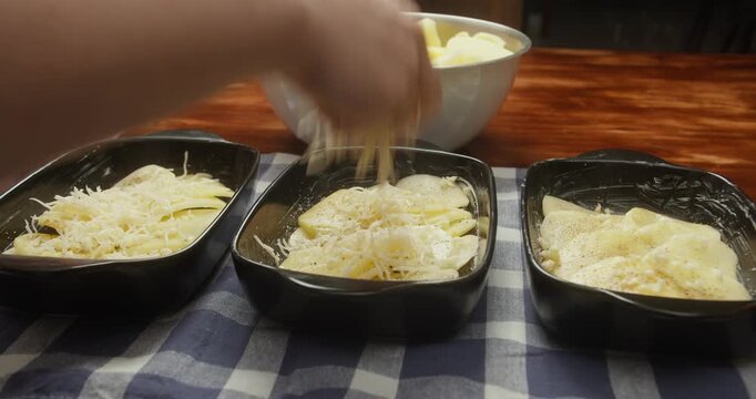 Chef sprinkles grated parmesan cheese over thinly sliced potatoes arranged in baking dishes. Close-up of food preparation, homemade potato casserole, rustic kitchen, natural light, traditional recipe
