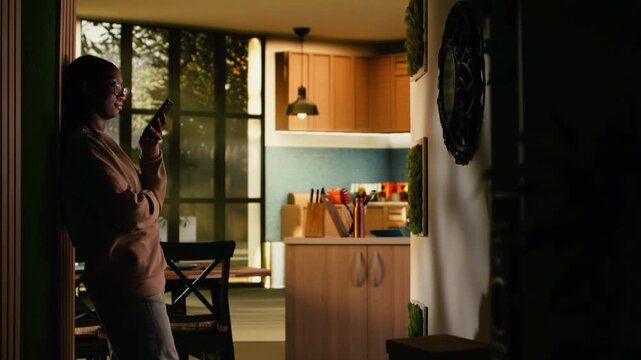 Young African American woman with phone sitting in the hallway in cozy ambiance, calm moment browsing mobile apps for web browsing. Warm lifestyle and casual screen time habits.