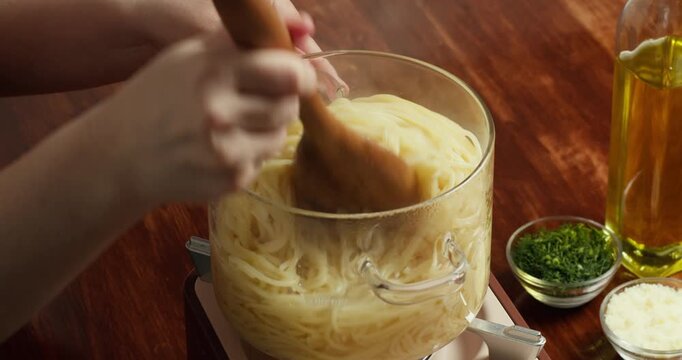 Close-up of hands mixing cooked spaghetti in pot with wooden spoon on wooden table. Homemade pasta preparation with olive oil, herbs and cheese. Traditional cooking process in kitchen, natural light.