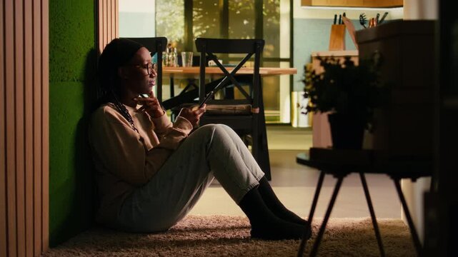 Young African American woman enjoying relaxation and browsing apps on phone, calm domestic life with warm interior for cozy lifestyle scene. Ambience focused on screen time and comfort indoor.