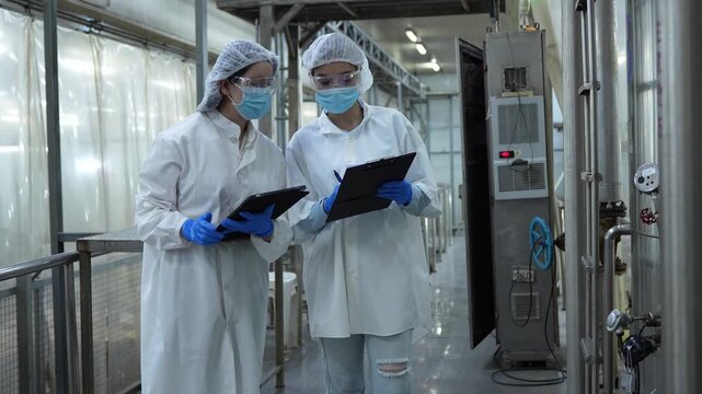 Asian female engineers using digital tablet and clipboard to inspect production machinery in a factory, Professional food safety auditors in lab coats monitoring manufacturing equipment
