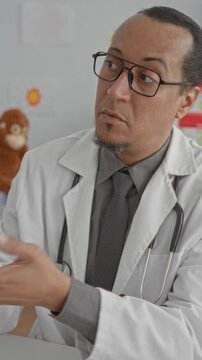 Man doctor wearing lab coat and stethoscope, gesturing with hands while seated at desk in a medical building; concern care.