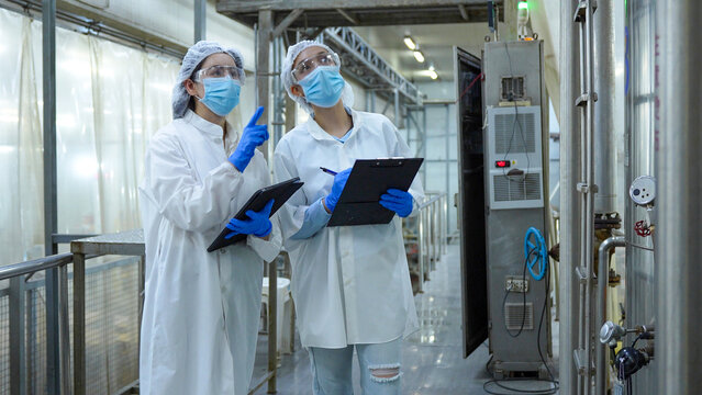 Female quality control inspectors in protective gear conducting audit in modern industrial plant, Professional food safety auditors wearing face masks and lab coats monitoring manufacturing equipment