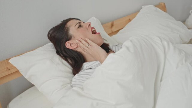 Woman on bed clutching jaw and hand, mouth open and grimacing in studio bedroom set with white bedding and wooden headboard; pain insomnia.