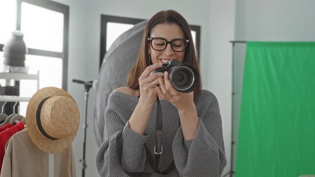 Young woman photographer holds camera to eye with finger on shutter, smiling amid green screen and clothing rack in studio; creative joy.