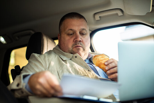 Businessman eating breakfast while working on laptop in parked car