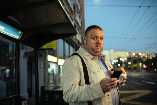 Middle-aged man waiting at city bus stop with coffee and smartphone at dawn