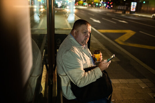 Man waiting at city bus stop at night with coffee