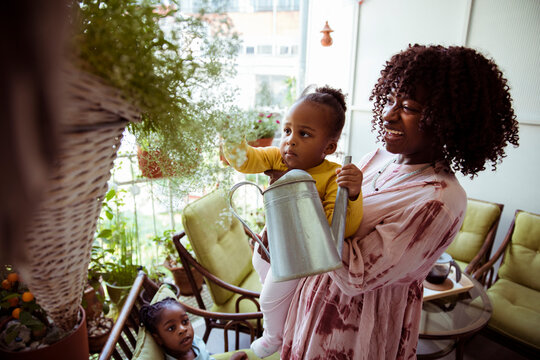 Mother helping toddler water plants on sunny balcony