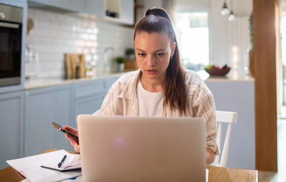 Young woman working on laptop with phone in home kitchen
