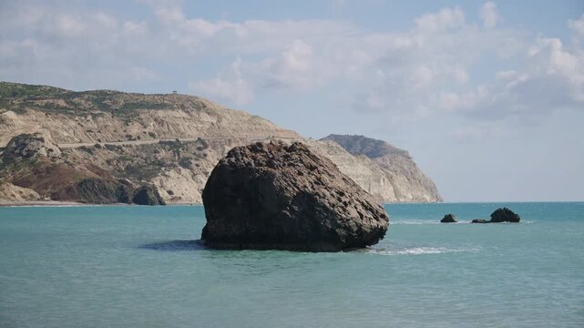 View of a large sea stack surrounded by calm water with coastal cliffs in the background near Aphrodite's Rock