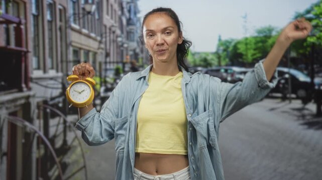 Young hispanic woman holding yellow alarm clock and pointing finger, bare midriff visible on a city street; urgency reminder.