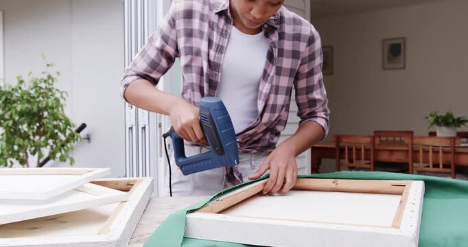 African American woman stretching green fabric and stapling frame with corded staple gun on porch