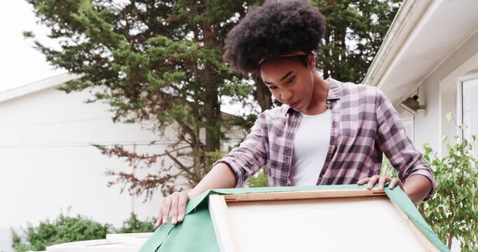 African American woman stretching green fabric on wooden frames at backyard table, stapler visible