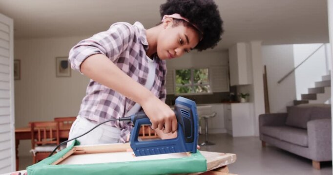 African American woman gripping blue stapler, leaning over frame and securing green fabric at home