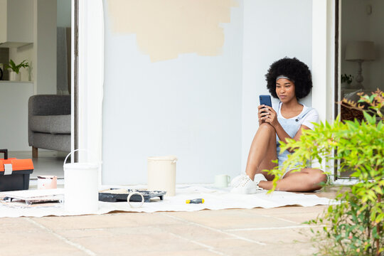 African American woman sitting at doorway, holding smartphone, paint cans on cloth, copy space
