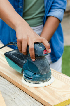 Woman sanding wood plank on outdoor bench, wearing green tee and denim, using teal sander