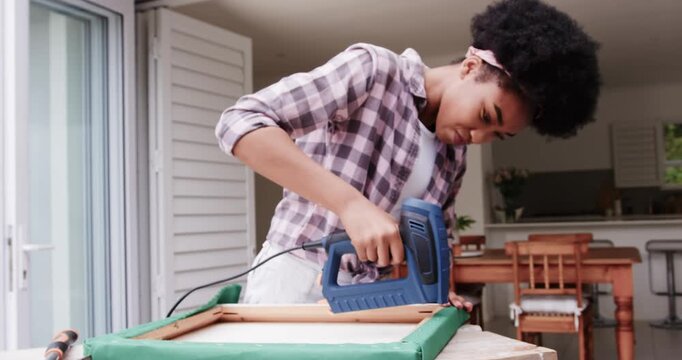 African woman placing frame, taking blue corded stapler, securing green fabric at table near door